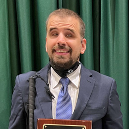 Corey stands in front of a dark green background. He is smiling and wearing a blue suit. He is holding his white cane. 