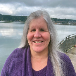 Kris stands in front of a body of water, smiling big, wearing a dark blue shirt. 