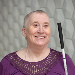 Marci Carpenter smiles with short hair, wearing a purple shirt. She is holding her long, white cane. 
