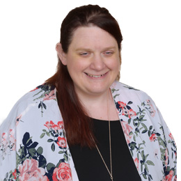 Chris Higley smiles with reddish brown hair in half up hairdo wearing a black top with an off-white cardigan with big pink and peach flowers with leaves Wearing a long gold necklace.